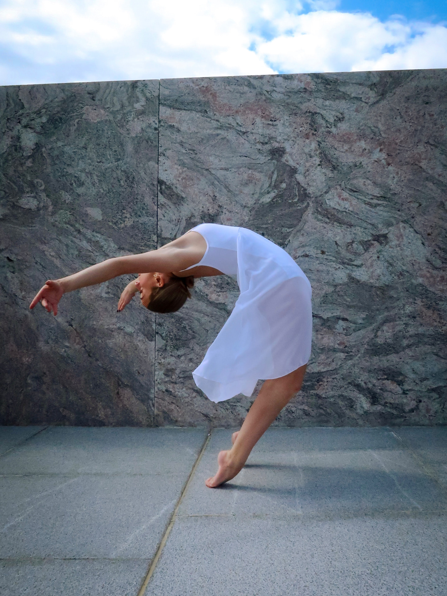 Nora in a backbend balance outdoors against a marble wall, one leg lifted and arms reaching behind her