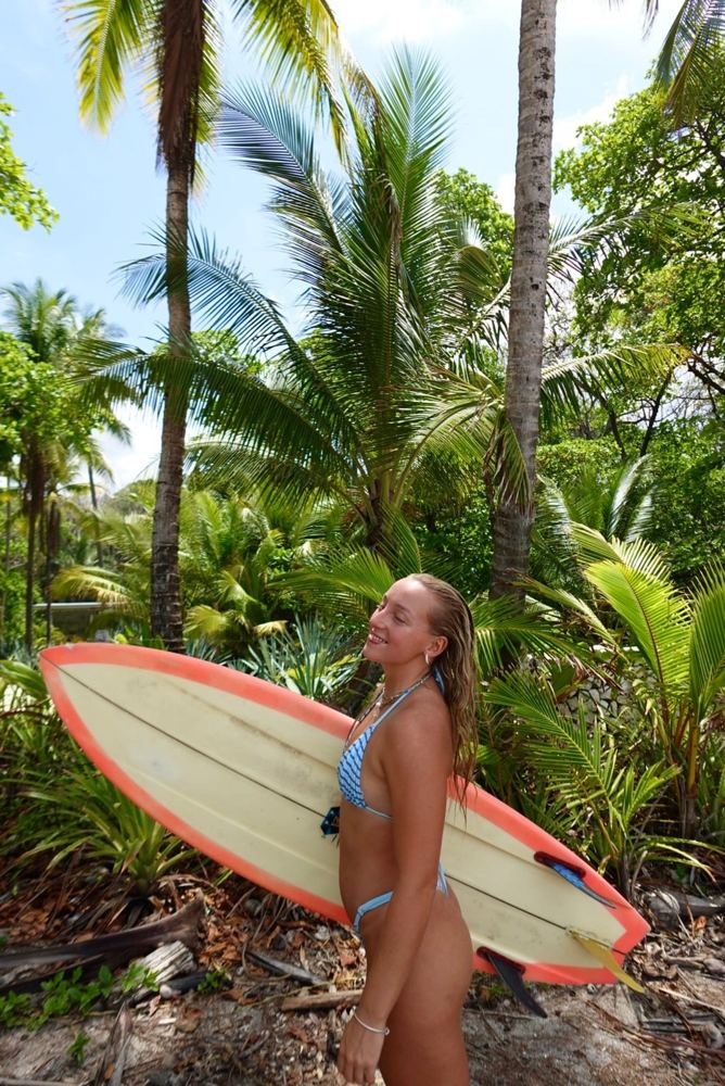 Nora holding a pink surfboard among palm trees